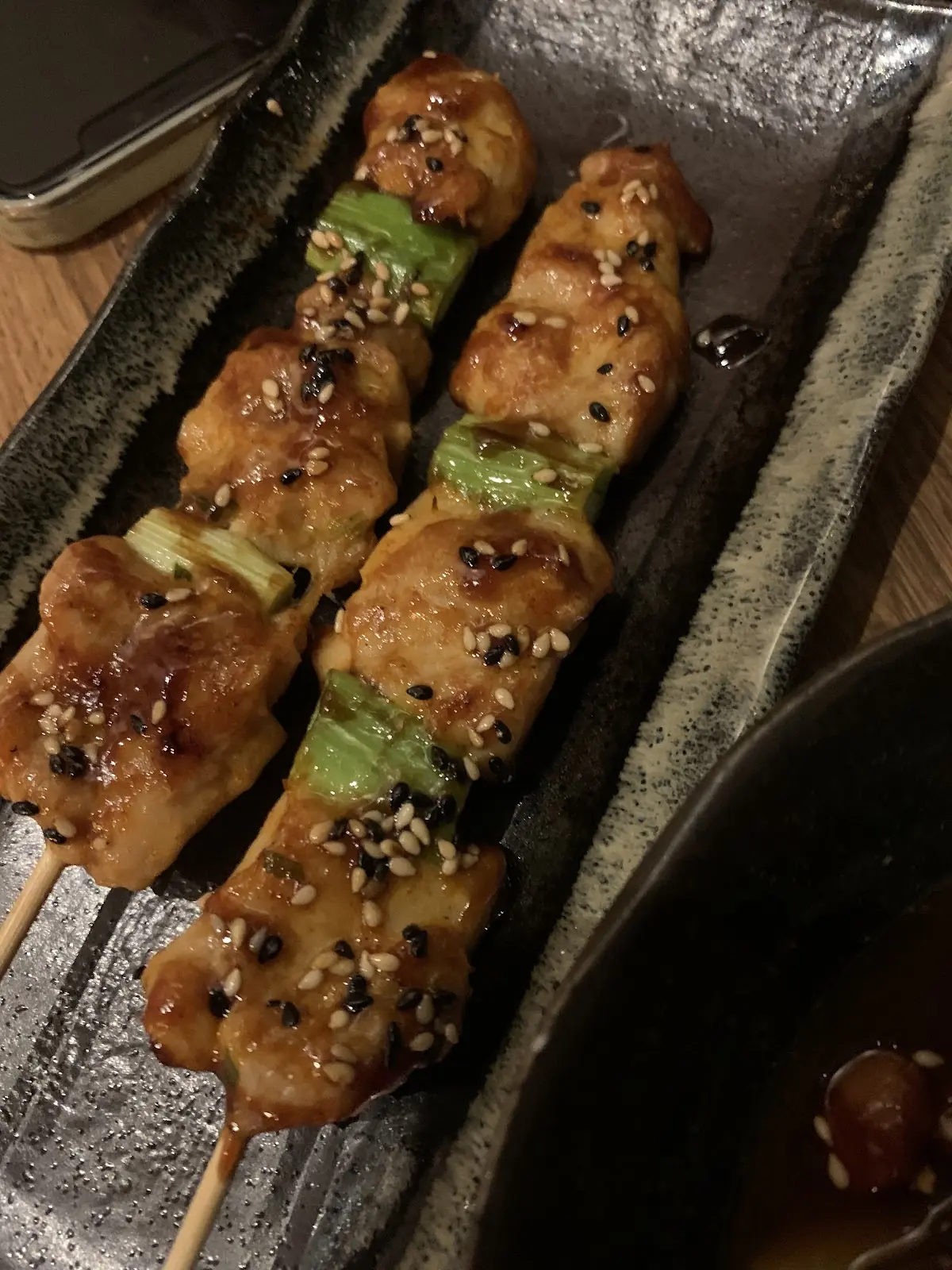 Two glazed chicken and spring onion yakitori skewers with sesame seeds on a long dark plate.