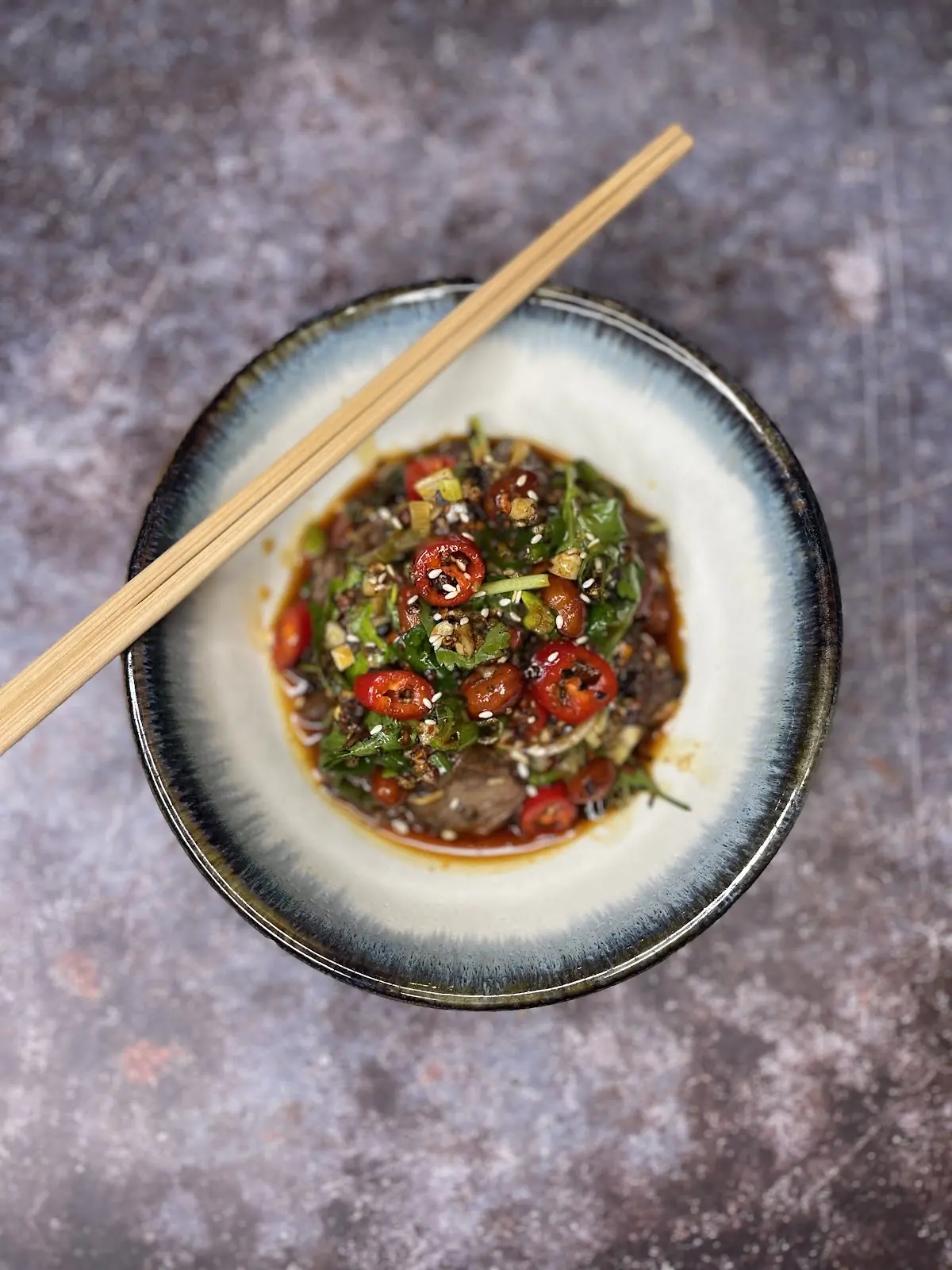 A shallow ceramic bowl of dark dipping sauce with sliced red chilli and spring onion, chopsticks resting across it.