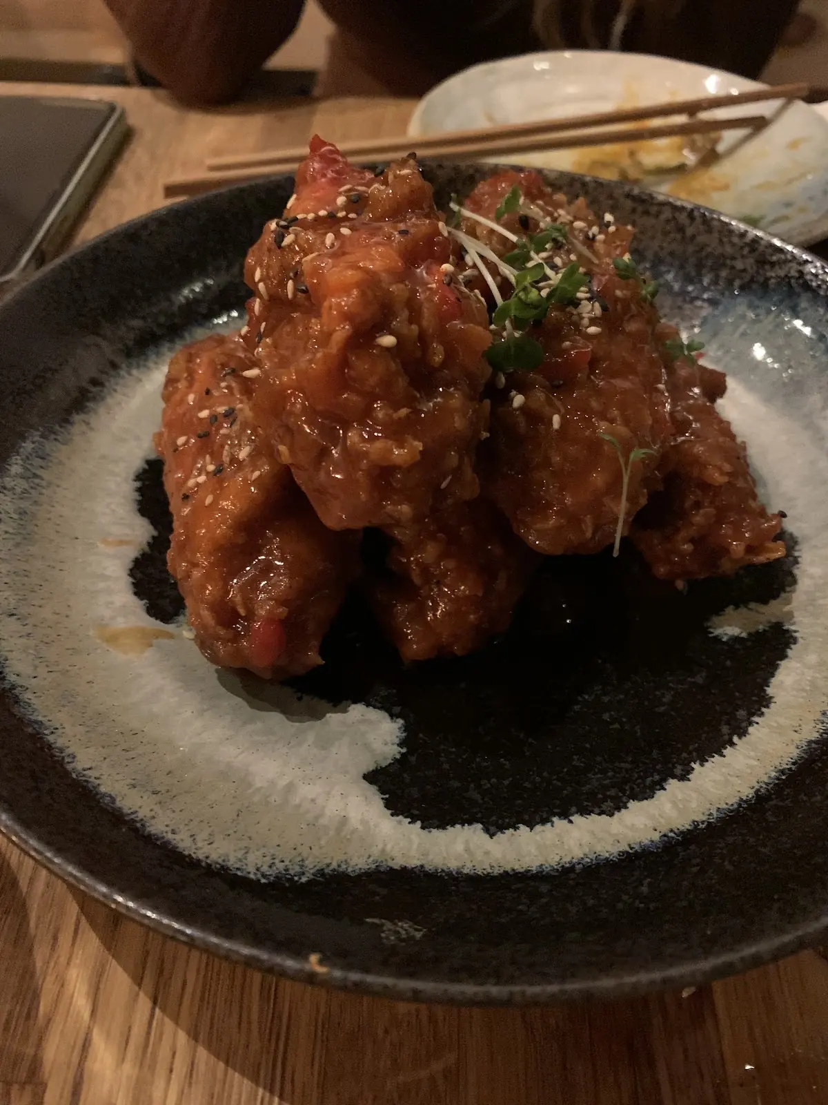 A pile of glossy red-sauced Korean fried chicken with sesame and microgreens on a black speckled plate.
