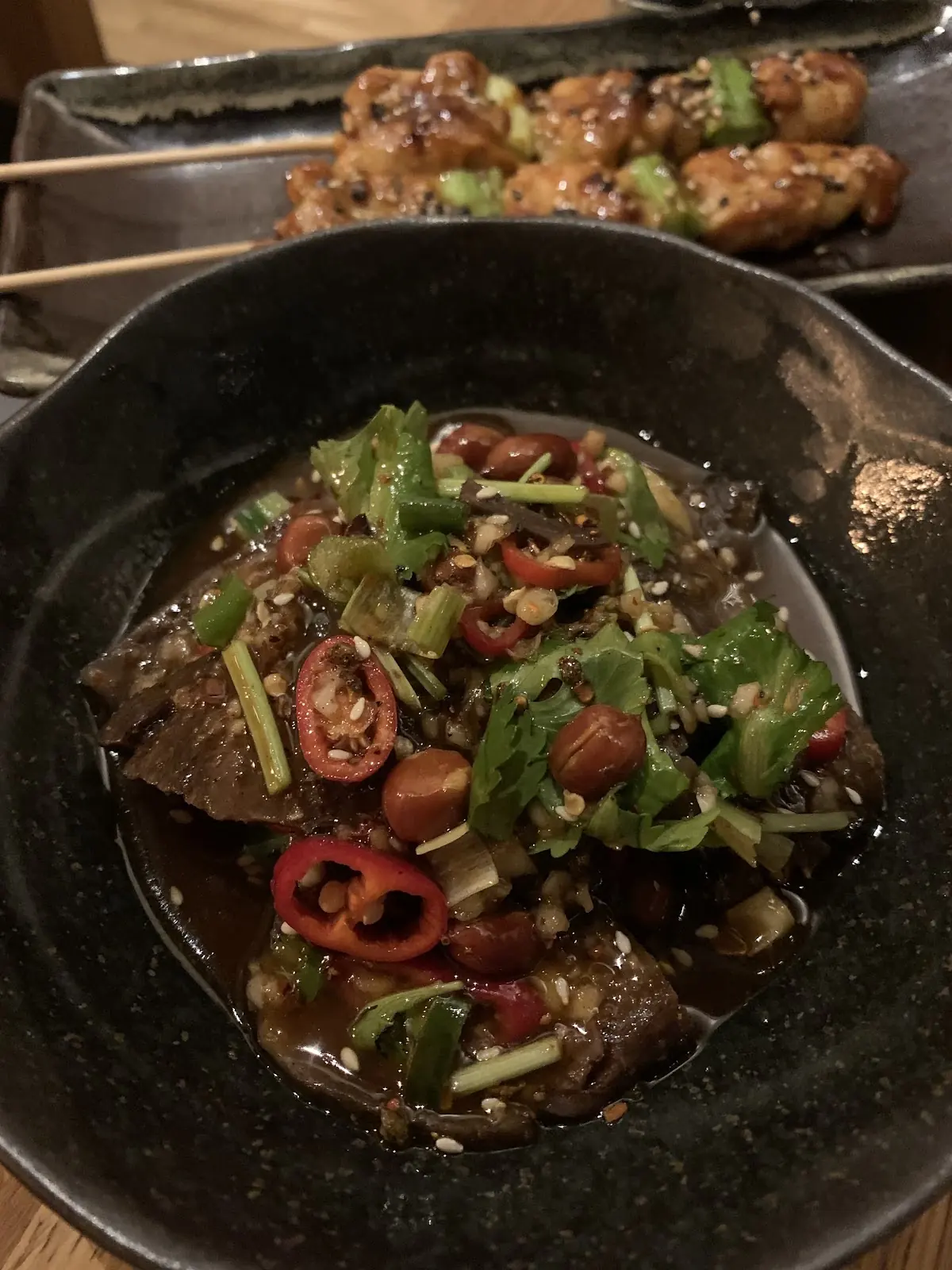 A dark bowl of braised meat topped with sliced red and green chillies and herbs, with grilled skewers behind it.
